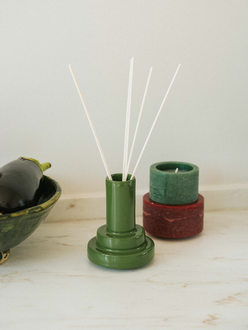 Un difusor de caña verde con un palo blanco se coloca sobre una superficie de mármol, junto a una berenjena en un cuenco verde y dos velas cilíndricas Stan Editions (una verde y otra roja).