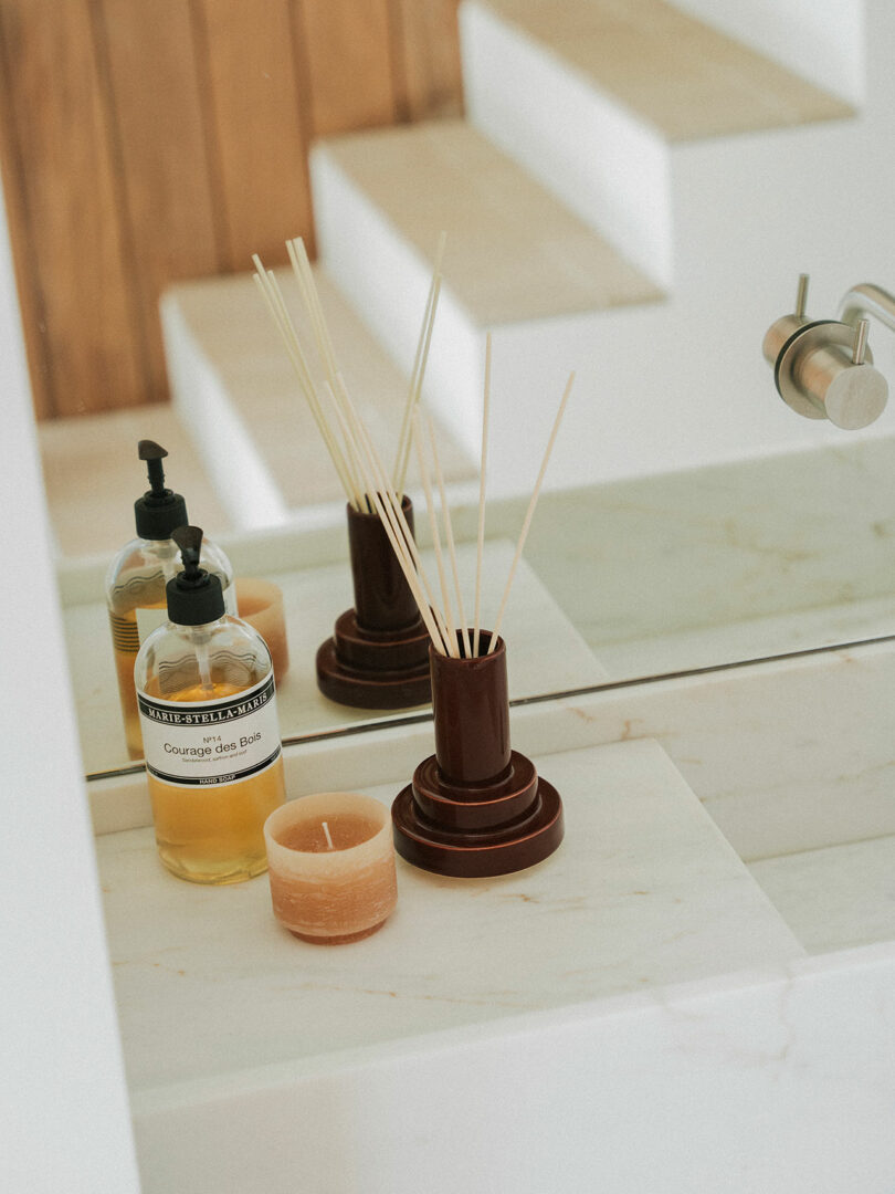 Un dispensador de jabón, un difusor de caña Stan Editions y pequeñas velas se encuentran sobre una encimera de mármol frente a un espejo cerca de una escalera blanca.