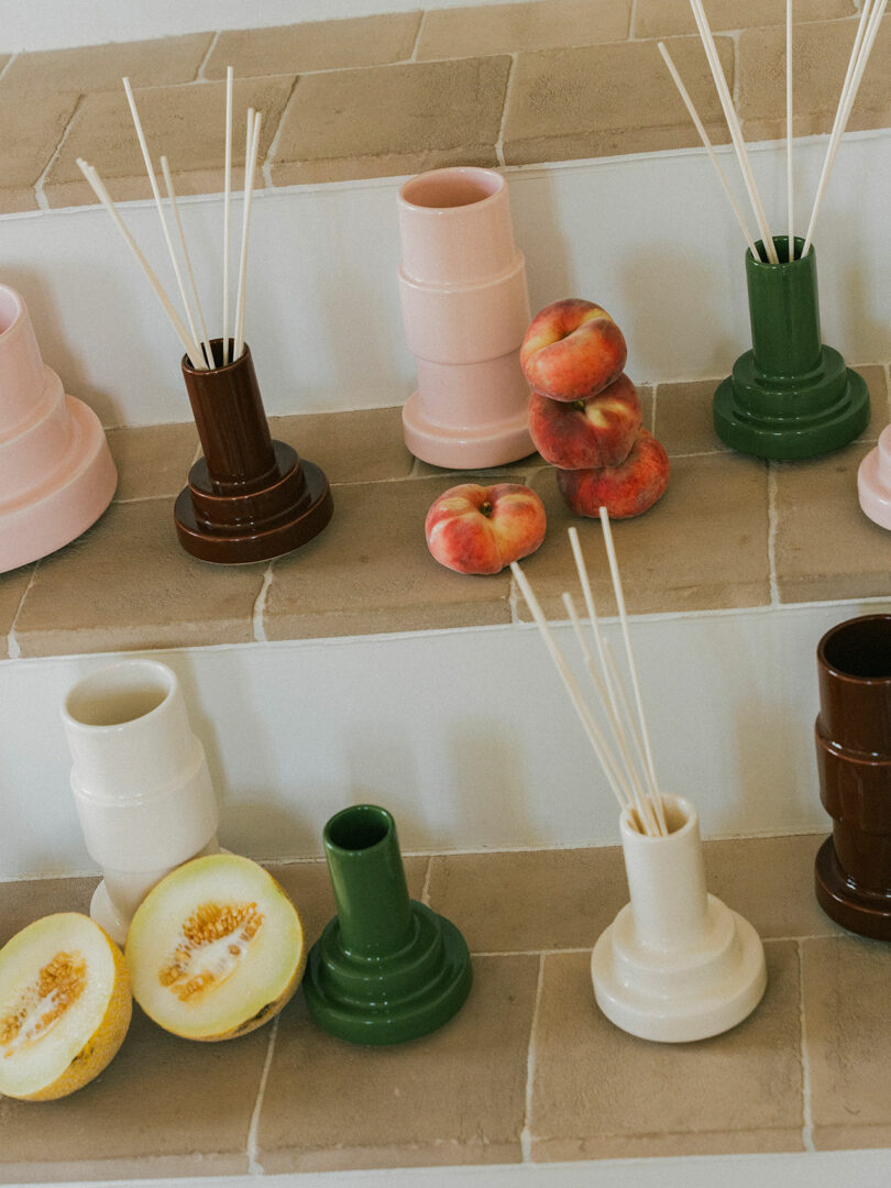 Jarrones de cerámica y difusores de caña de Stan Editions en varios colores se alinean en los escalones de azulejos junto con rodajas de melocotones y pepinos.