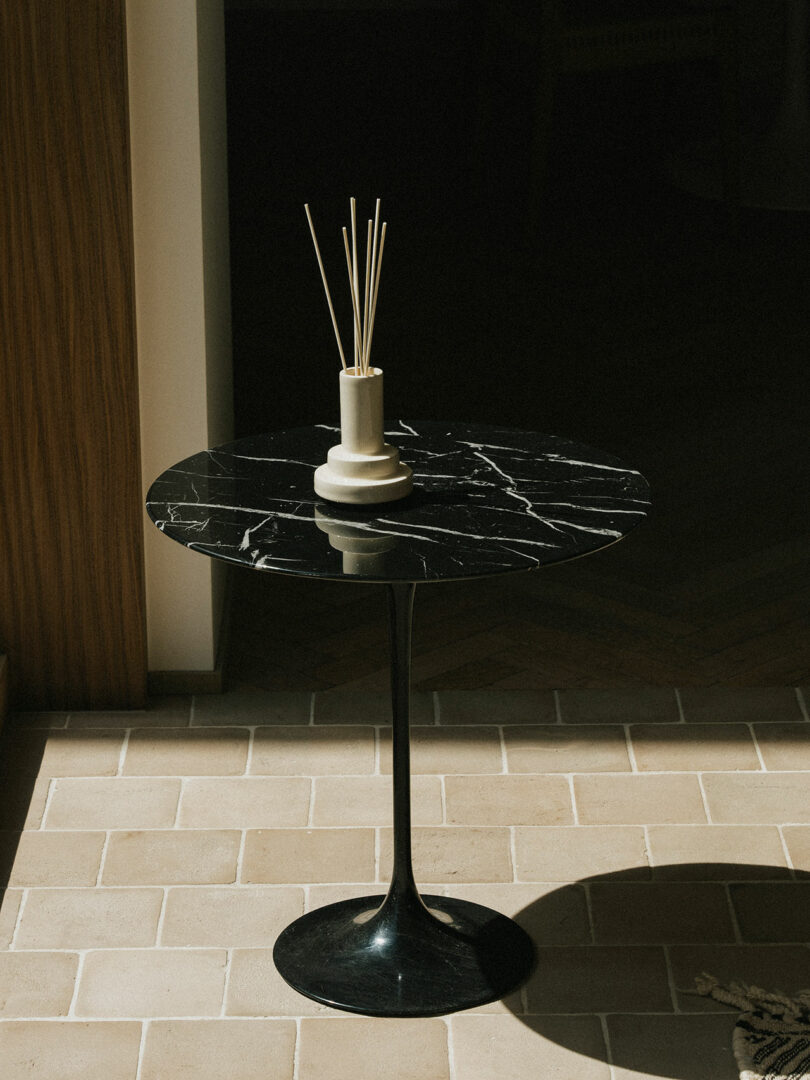 Una pequeña mesa auxiliar redonda de mármol negro Stan Editions, rematada con un difusor y varillas de caña, se encuentra sobre el suelo de baldosas de una habitación soleada.