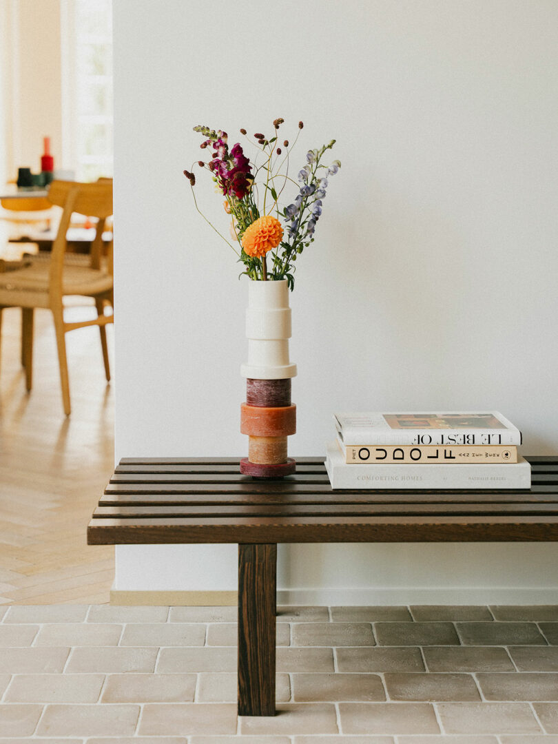 Un banco de madera descansa contra una pared blanca, sobre la cual hay un moderno jarrón estarcido con flores de colores y dos libros. Al fondo se ve la zona del comedor.