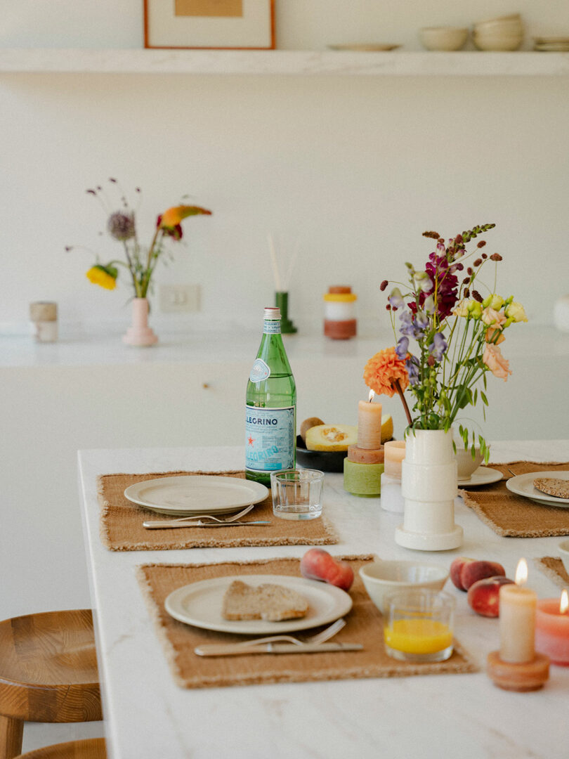 La mesa del desayuno está dispuesta con platos, pan, fruta, jugo, una botella de refresco, velas Stan Editions y un jarrón de flores; Se pueden ver más decoraciones en los estantes del fondo.