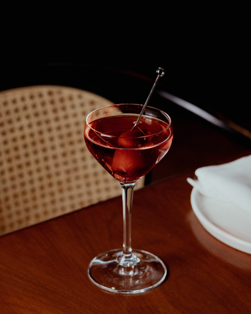 A cocktail in a stemmed glass with a cherry garnish on a metal pick sits on a wooden table next to a white napkin and a chair with a woven back.