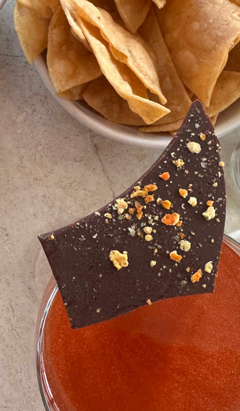 A piece of dark chocolate with seasoning is held above a glass of red liquid, with a bowl of tortilla chips in the background.