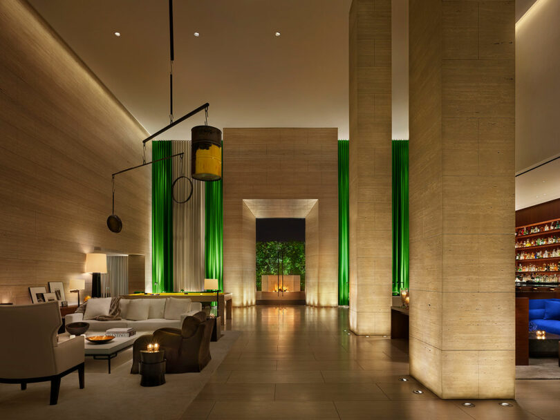 Modern hotel lobby with high ceilings, neutral-toned walls, contemporary furniture, green accent lighting, and a well-lit bar area in the background.
