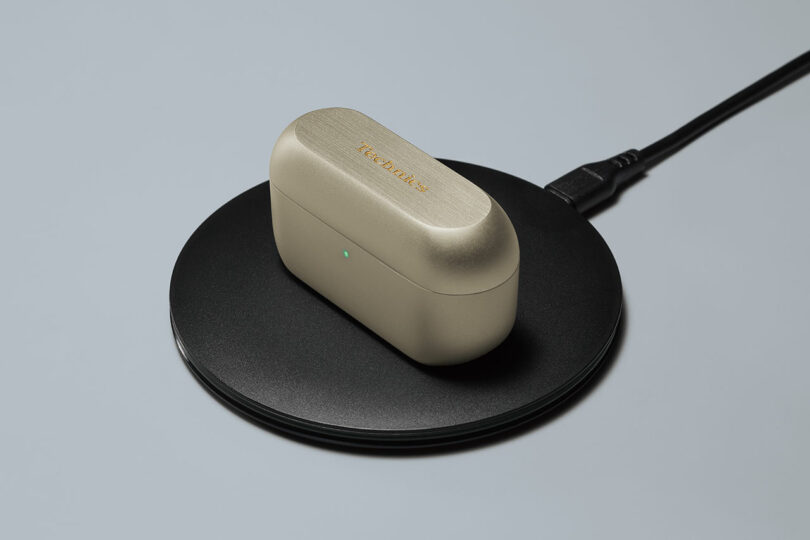 A beige wireless earbud case with a green indicator light sits on a black wireless charging pad connected by a cable.