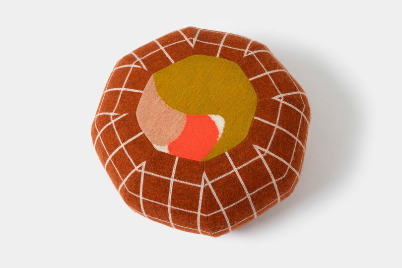 Round, brown SCP cushion with a white grid pattern and abstract shapes in yellow, red, and beige on the top, photographed against a white background.
