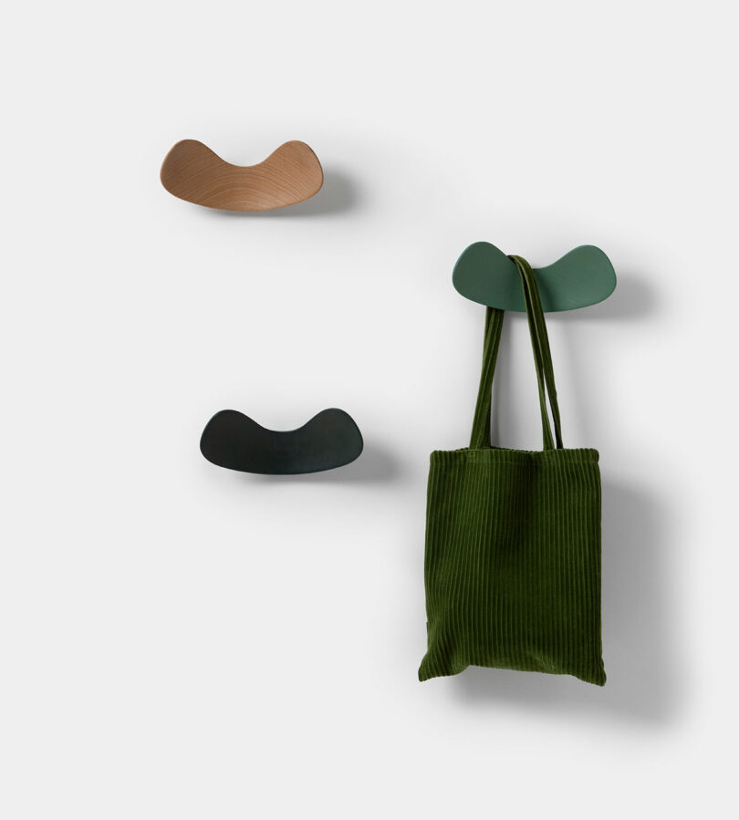 Three curved SCP wall hooks in green, brown, and black hold a green corduroy bag against a plain light background.