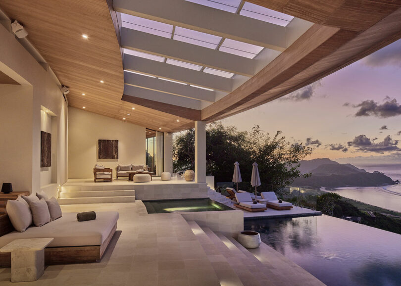 Modern open-air lounge with poolside seating, infinity pool, and a view of mountains and the ocean at sunset.