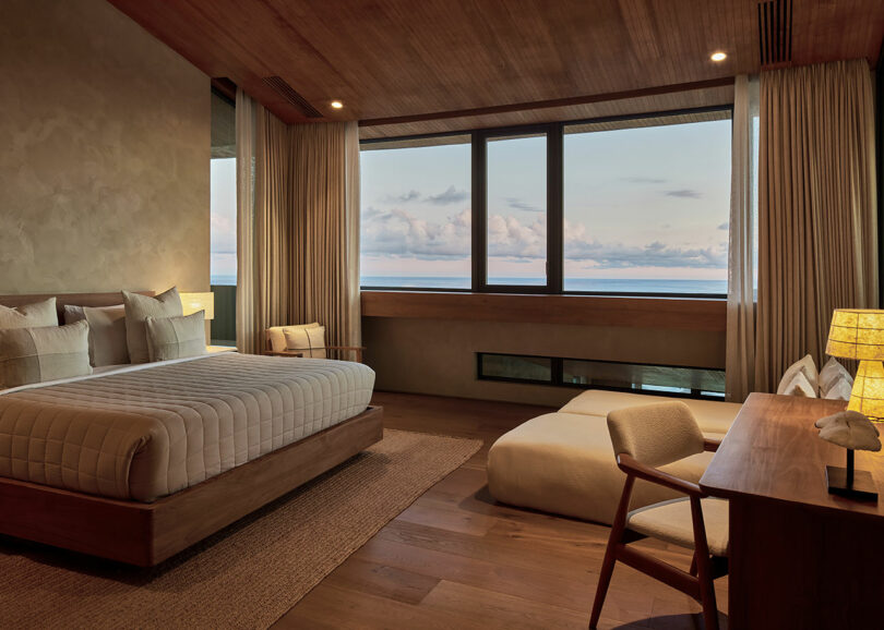 Modern bedroom with a large bed, desk, and lounge chair, featuring wood finishes, neutral tones, and large windows overlooking the ocean at sunset.