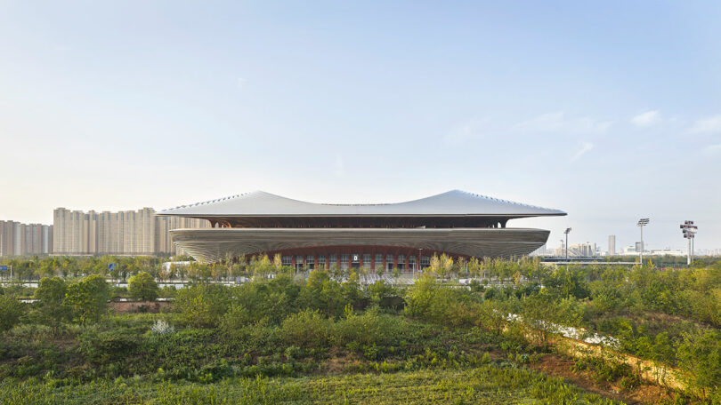 A modern stadium with a curved roof stands amid greenery, with high-rise buildings visible in the background under a clear sky.
