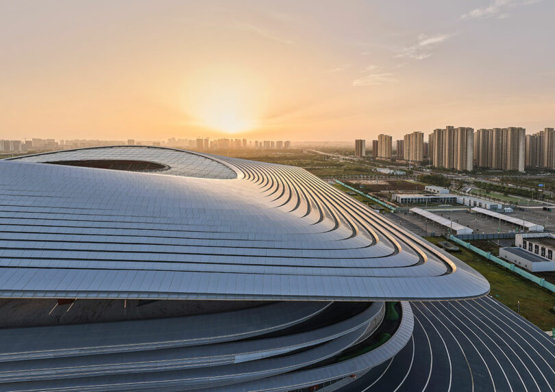 A modern, curved building with metallic surfaces is shown at sunset, with high-rise apartments and city infrastructure visible in the background.