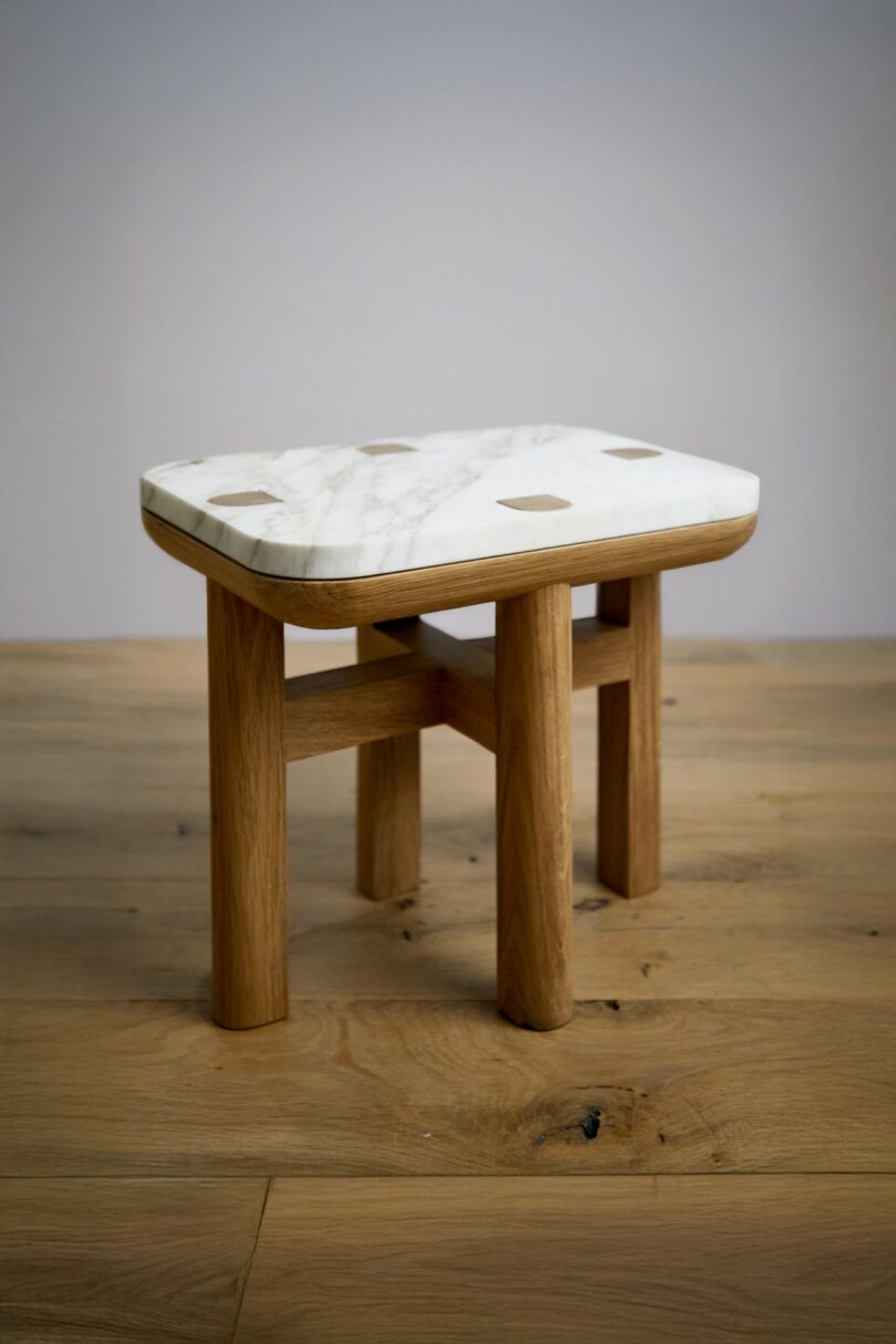 A small wooden stool with a square marble seat and four legs, placed on a wooden floor against a plain background
