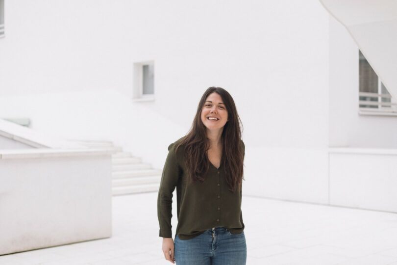 A woman with long dark hair, wearing a green shirt and jeans, stands and smiles in a bright, minimalist outdoor setting with white walls and stairs