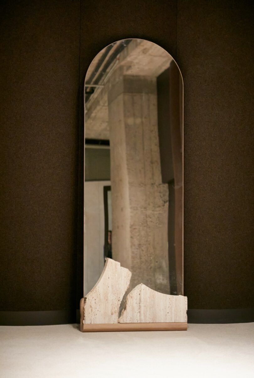 A tall, arched mirror with a stone base and an irregular, jagged lower edge stands against a dark wall, reflecting a concrete pillar and ceiling
