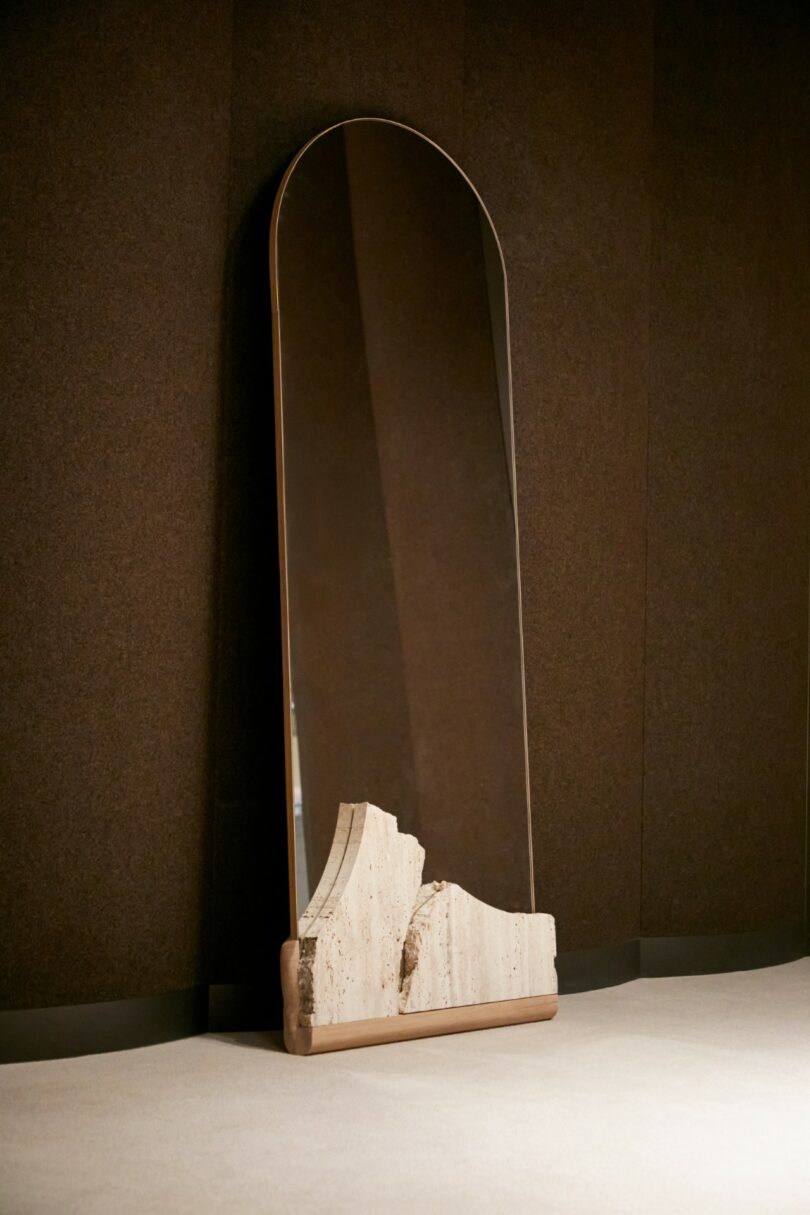 A tall, arched mirror with a sculpted stone base stands against a dark brown wall on a light-colored floor