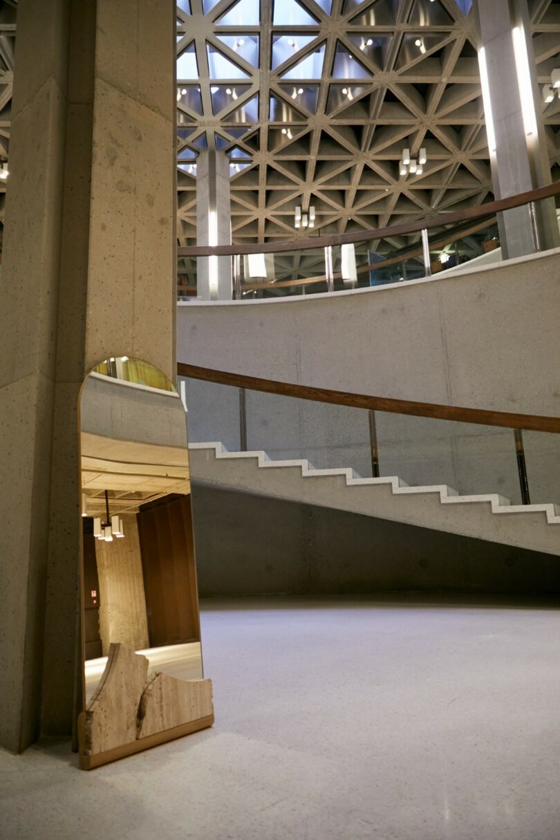 A broken mirror rests against a concrete pillar in a modern building with geometric ceiling patterns and a curved staircase