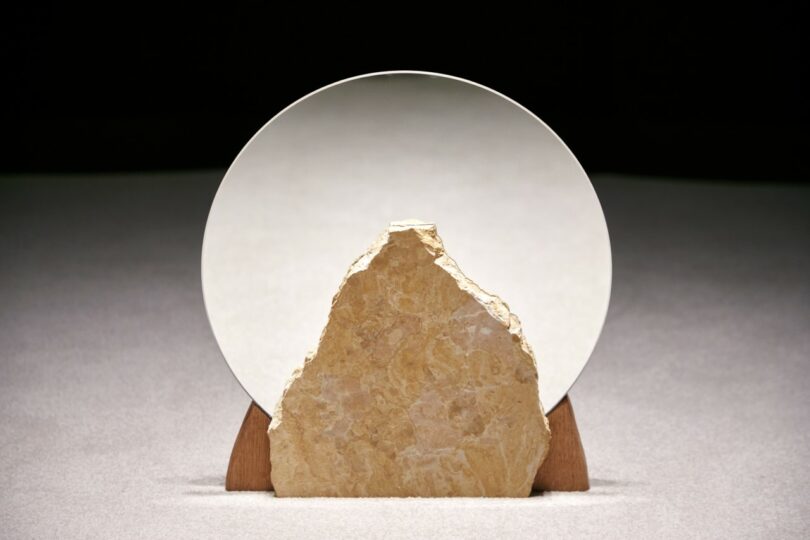 A rough, triangular stone is positioned in front of a circular mirror on a wooden stand, casting a partial reflection against a neutral background