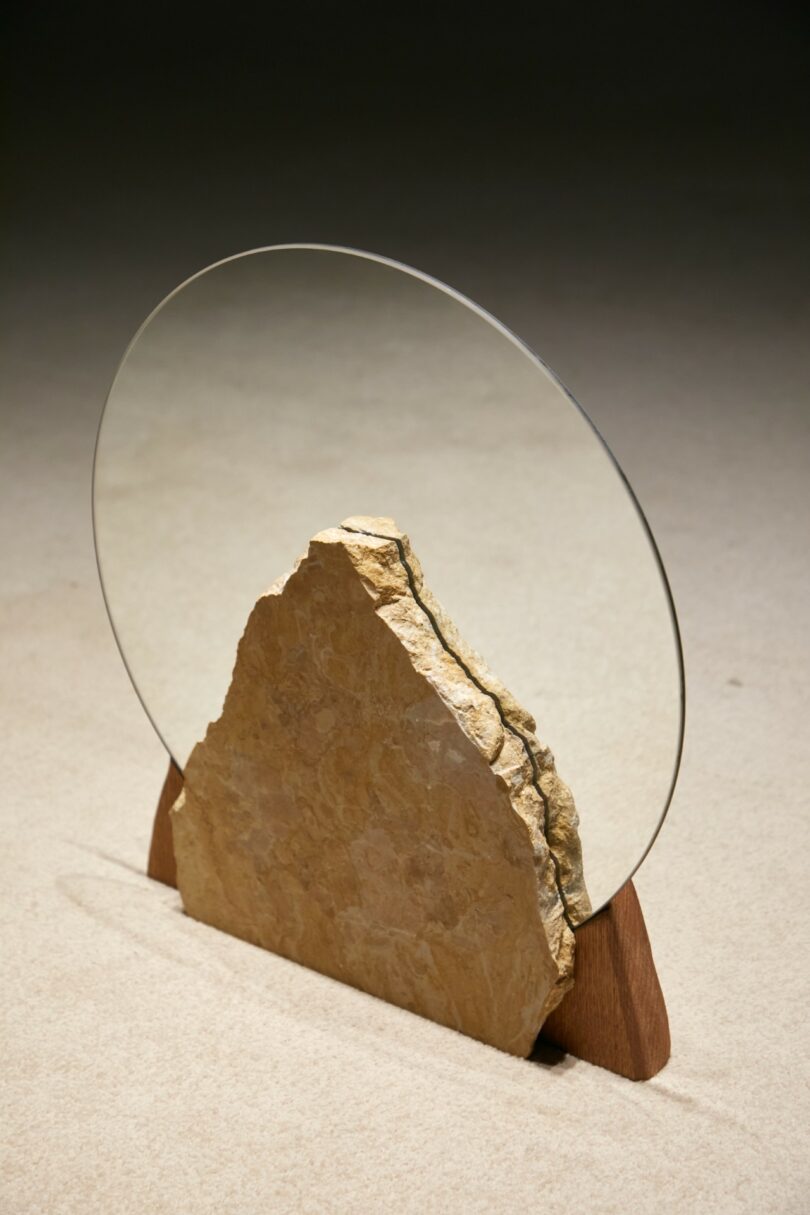 A large stone slab and a round glass pane are vertically positioned together, supported by two wooden stands, on a neutral background