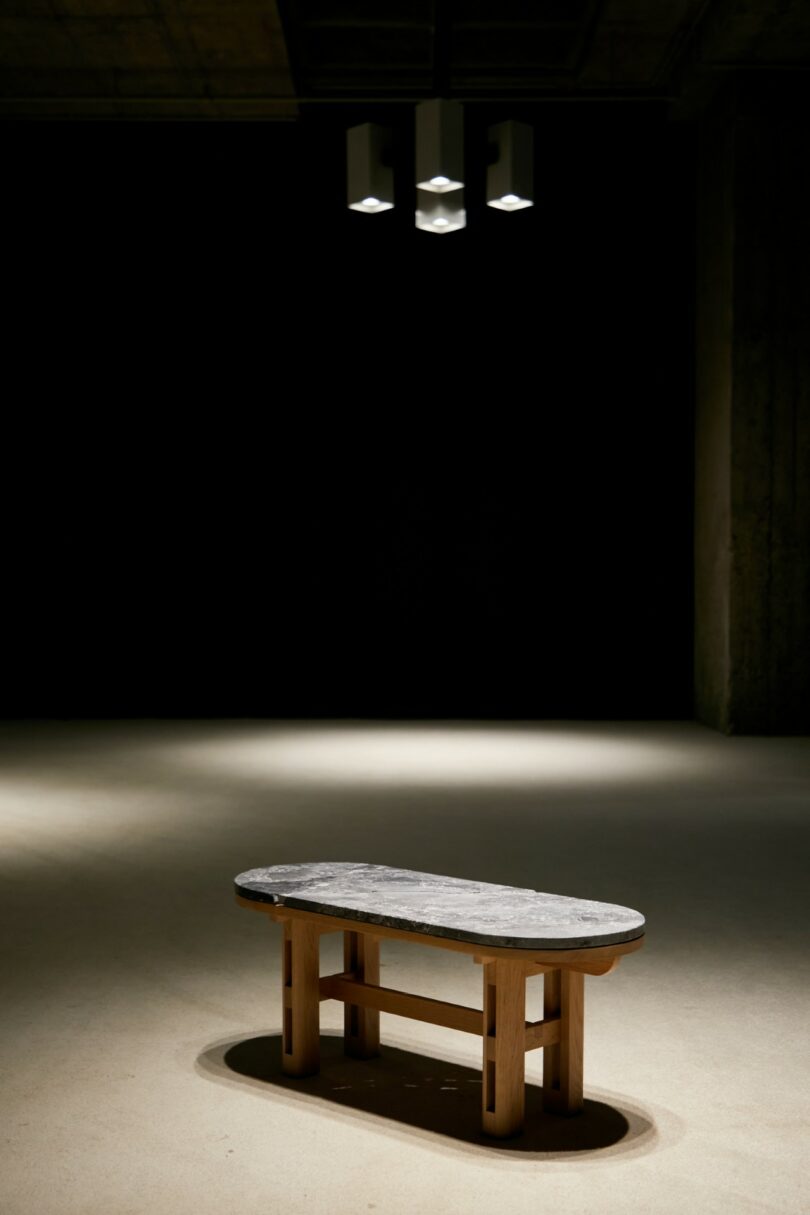 A stone-topped wooden bench is positioned under a ceiling light in an otherwise dark, empty room with concrete floors and walls