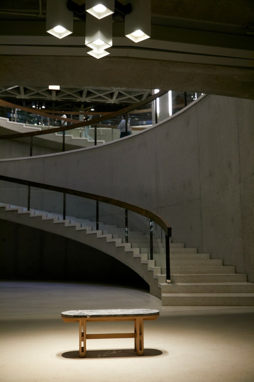 A modern, empty interior features a curved staircase, concrete walls, a round table in the center, and geometric ceiling lights above