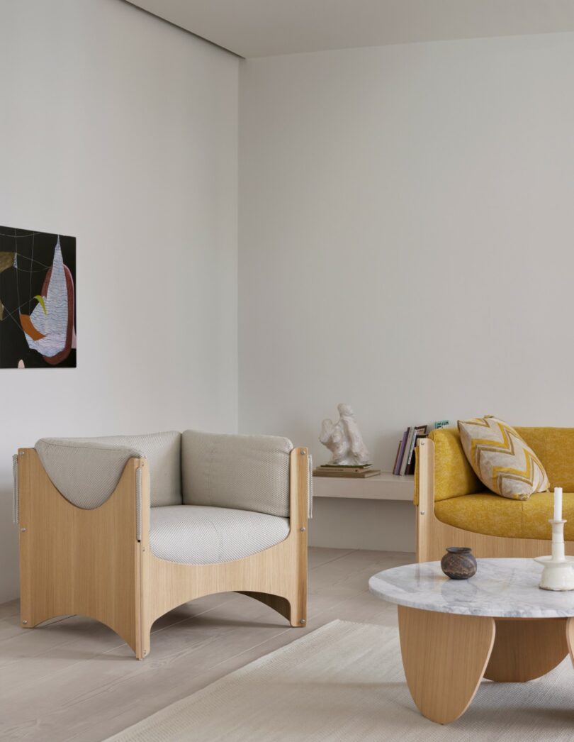 Salón moderno con sillón de madera clara con cojines claros, sofá amarillo, mesa baja redonda de mármol y una pequeña repisa con libros y adornos en una pared blanca