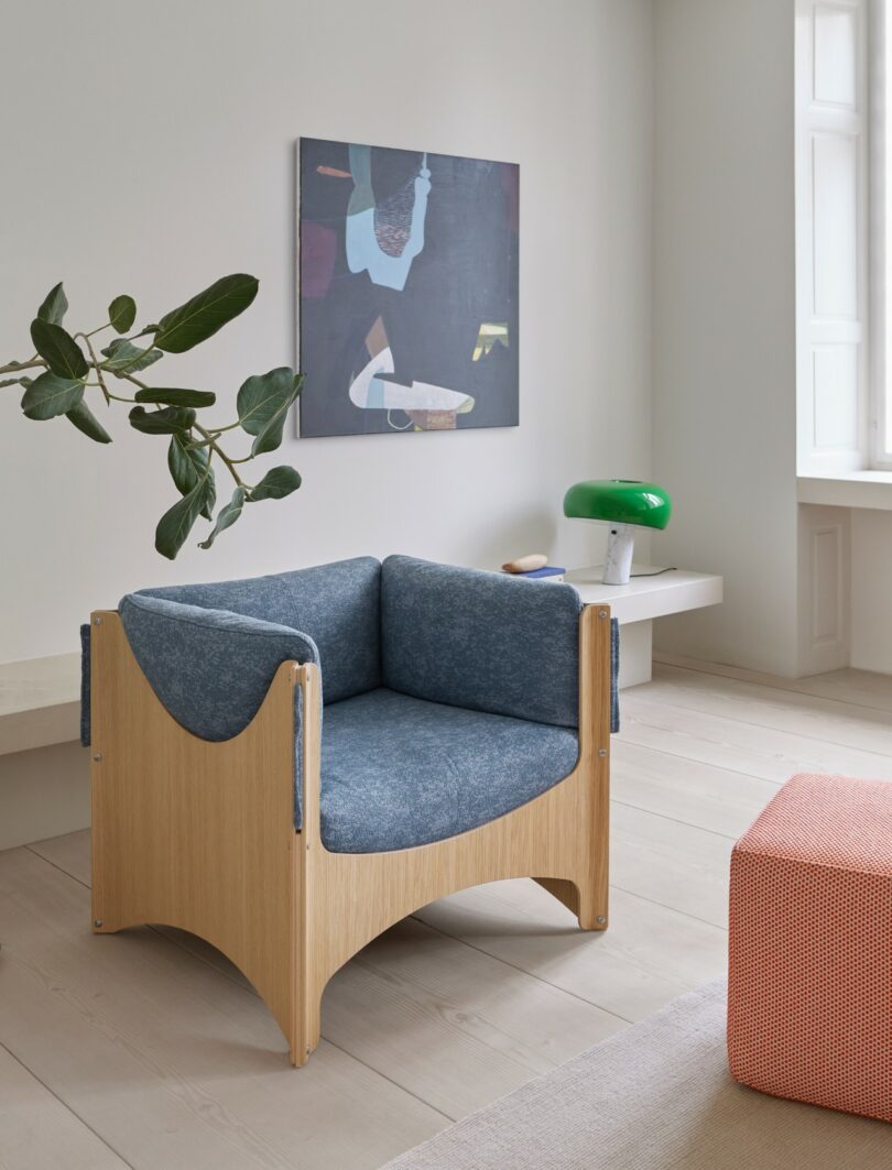 Un sillón moderno con tapizado azul y estructura de madera clara se encuentra en una habitación minimalista con arte en la pared, una lámpara verde sobre un estante blanco, una planta y un taburete rojo.