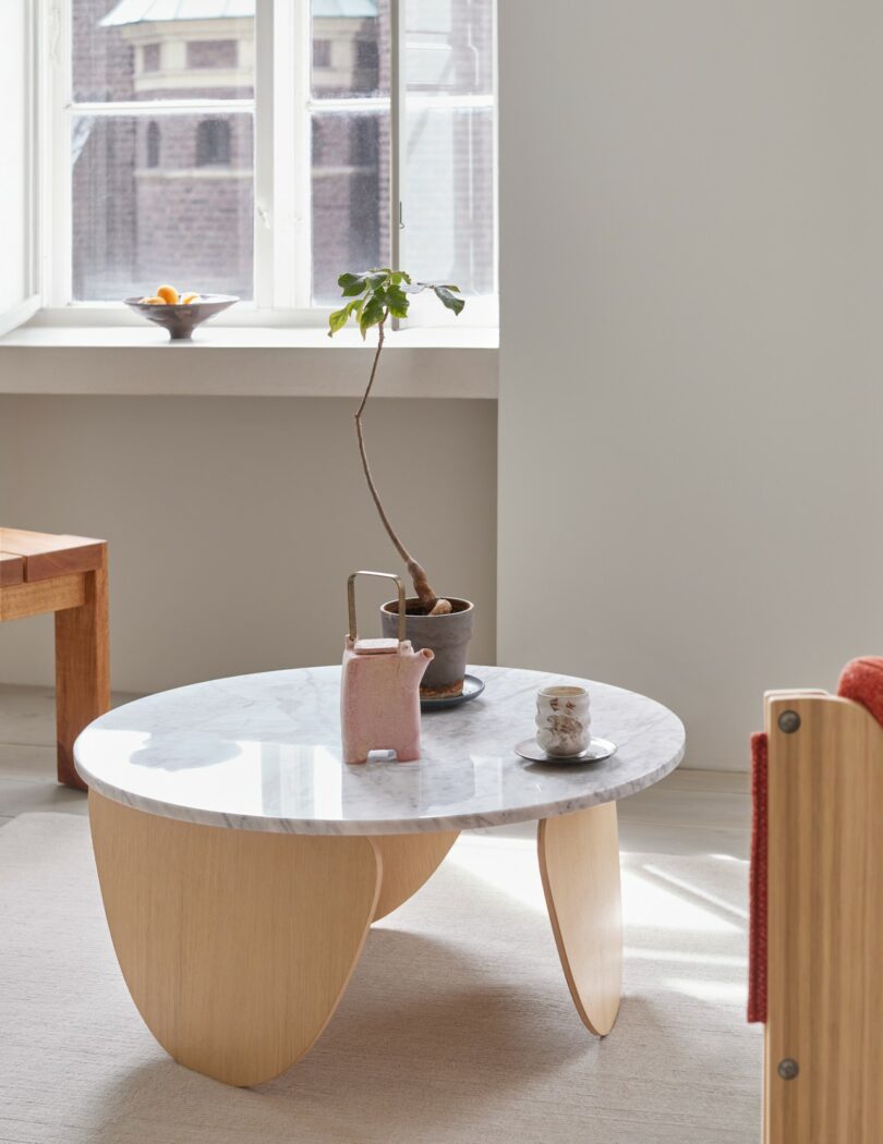 Una pequeña mesa de centro redonda de mármol con patas de madera, una maceta, una jarra de cerámica y tazas, cerca de una ventana por la que entra la luz del sol