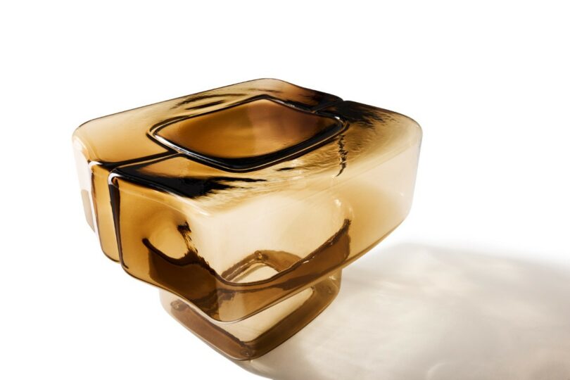 A geometric, amber-colored glass sculpture with smooth edges and angular lines, displayed on a white background with a subtle shadow