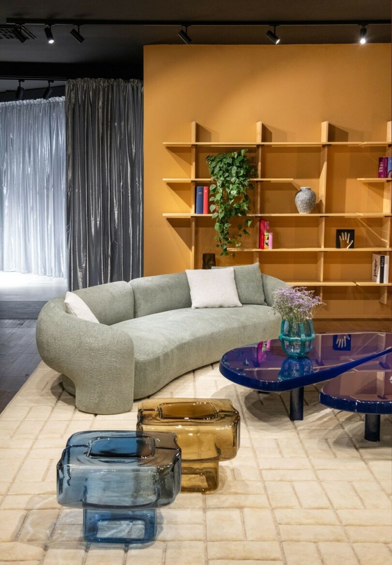 A modern living room with a light green curved sofa, a blue coffee table, glass stools, a beige rug, and a yellow wall featuring shelves with books, decor, and a hanging plant