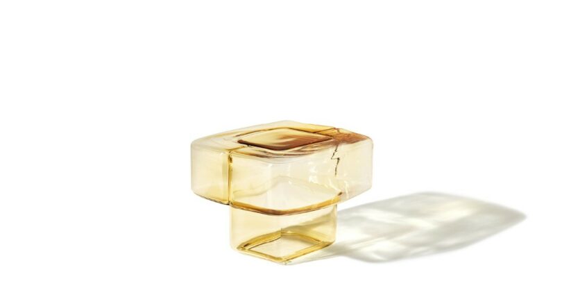 A translucent amber glass object with a square top and a hollow rectangular base, casting a soft shadow on a white background