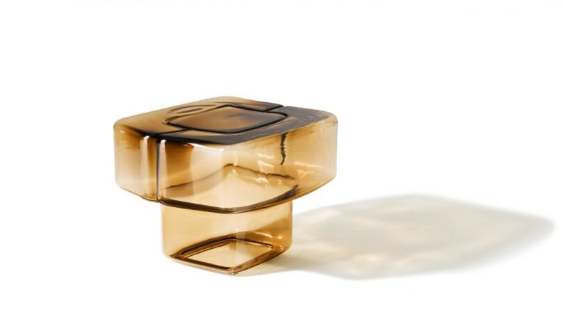 A cube-shaped amber glass table with rounded edges and a hollow base, casting a light shadow on a white background