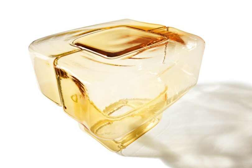 A sculptural amber-colored glass object with smooth, geometric surfaces and rounded edges, casting a faint shadow on a white background