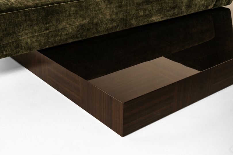 Primer plano de una plataforma de madera oscura y lisa que sostiene la base de un sofá tapizado de color verde oliva, sobre un fondo blanco