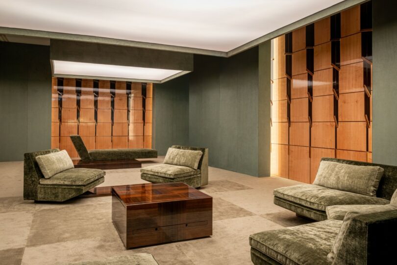 Salón moderno con sillas de terciopelo verde, mesa de café de madera y paredes con paneles de madera bajo grandes luces empotradas en el techo