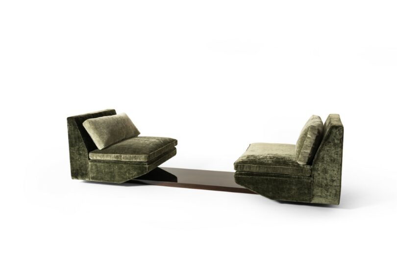 Dos sillones de terciopelo verde uno frente al otro, con bases conectadas entre sí por una mesa baja de madera oscura, sobre un fondo blanco puro