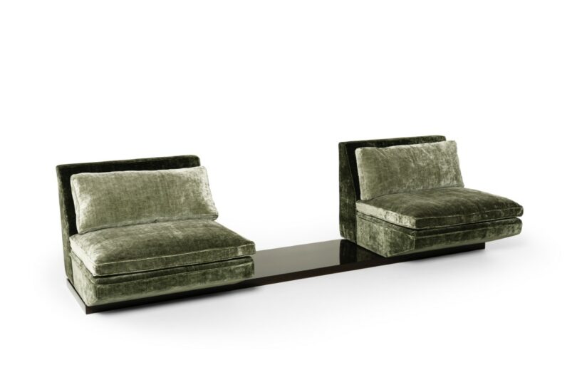 Dos sillones adyacentes de terciopelo verde con bases conectadas por una mesa baja de madera oscura sobre un fondo blanco puro