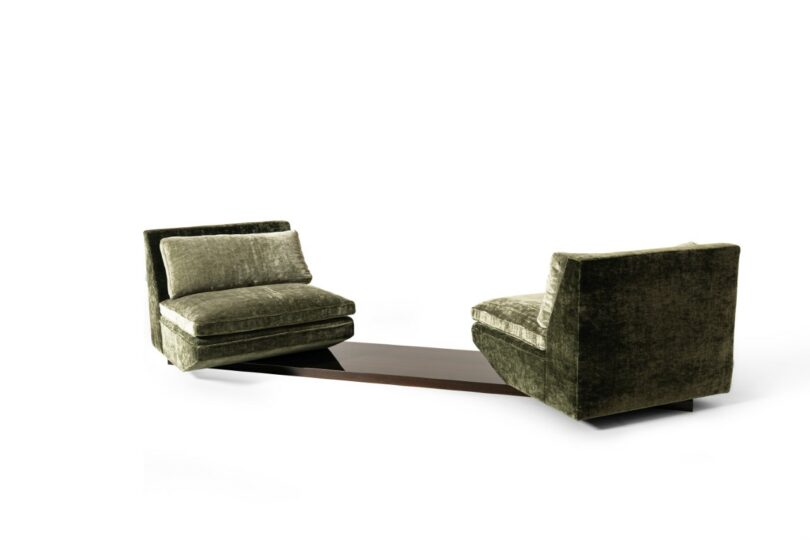 Dos sillones adyacentes de terciopelo verde con bases conectadas por una mesa baja de madera oscura sobre un fondo blanco puro