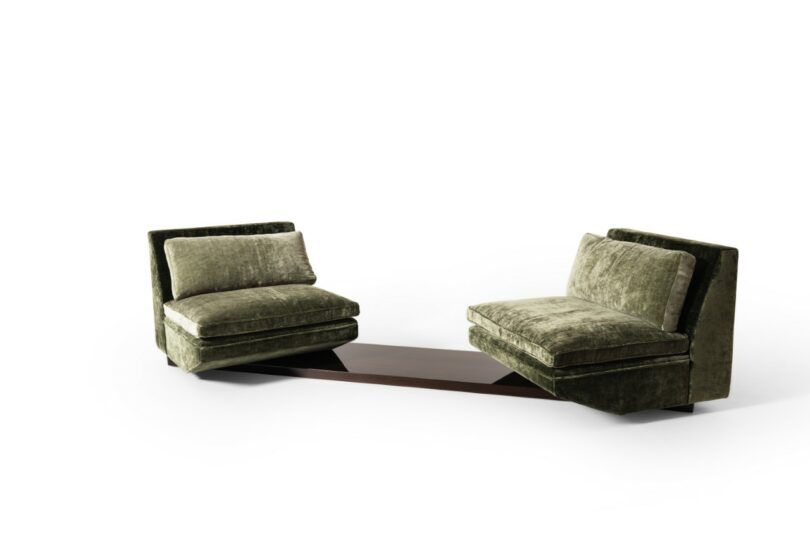 Dos sillones adyacentes de terciopelo verde con bases conectadas por una mesa baja de madera oscura sobre un fondo blanco puro