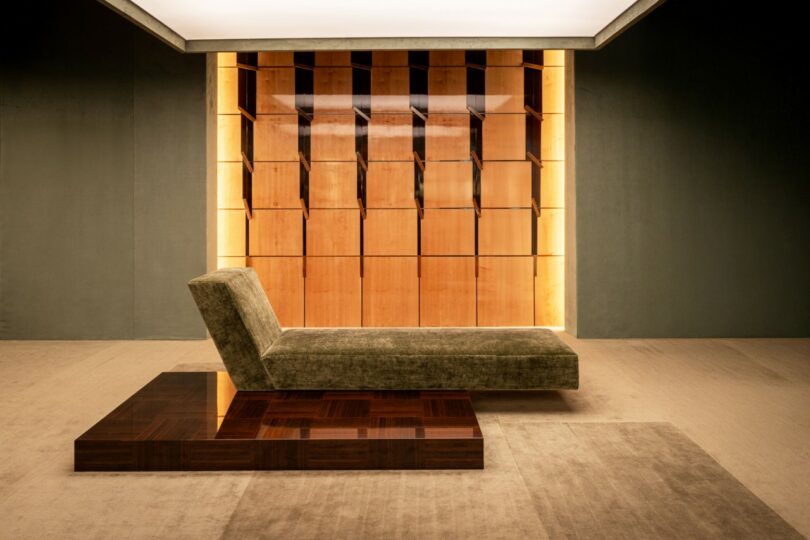 Un moderno sillón con cojines verde-marrón se asienta sobre una plataforma de madera frente a una pared de madera retroiluminada con paneles cuadrados