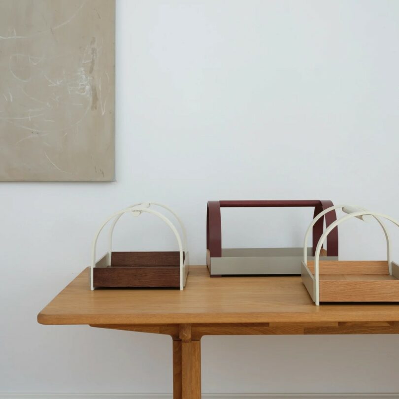 A wooden table with three rectangular trays featuring arched handles, placed beneath an abstract beige painting on a white wall