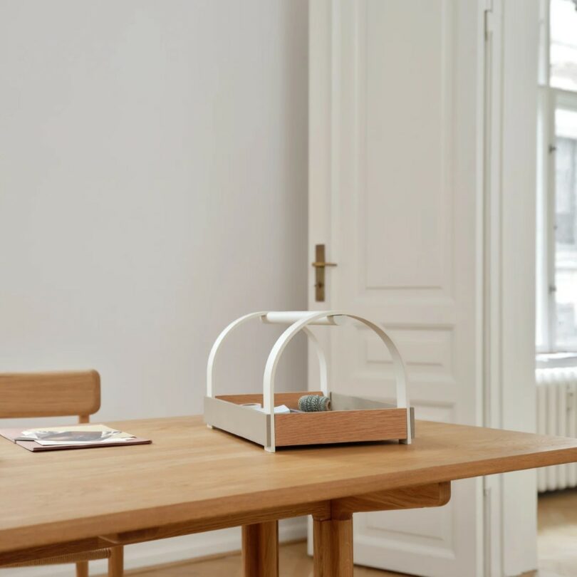 A wooden table with a wooden and white metal tray, a rolled-up cloth, and papers, set in a minimalistic room with white walls and a door