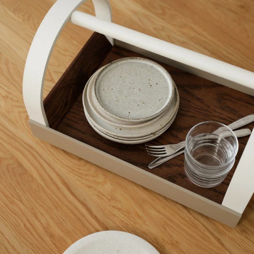 A wooden tray with a white handle holds stacked ceramic plates, a glass of water, a fork, and a spoon on a wooden table