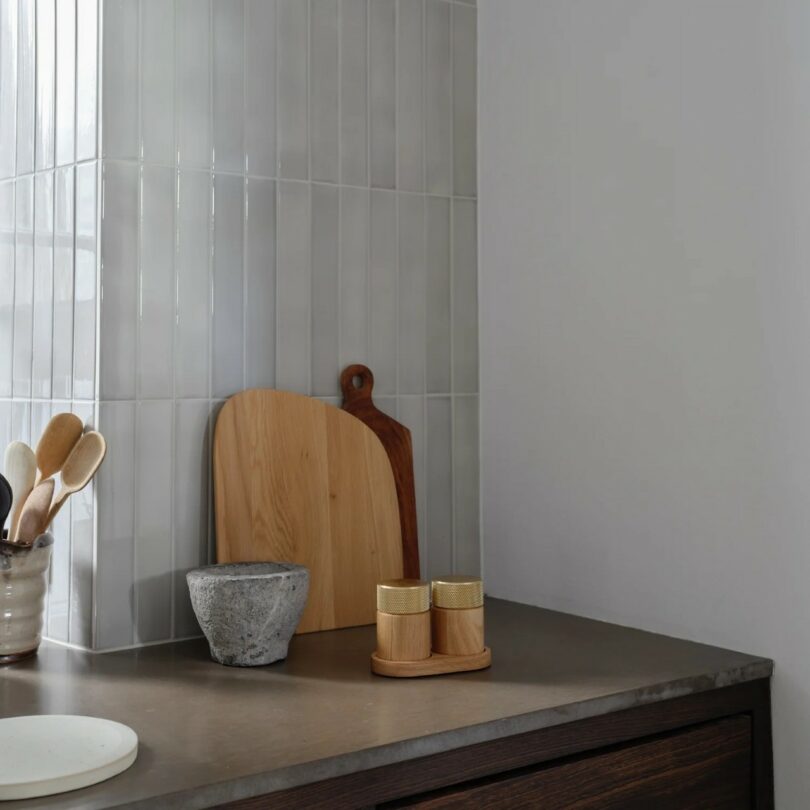 A modern kitchen countertop with two cutting boards, a stone mortar, spice jars, and a jar holding wooden utensils, set against light vertical tiles