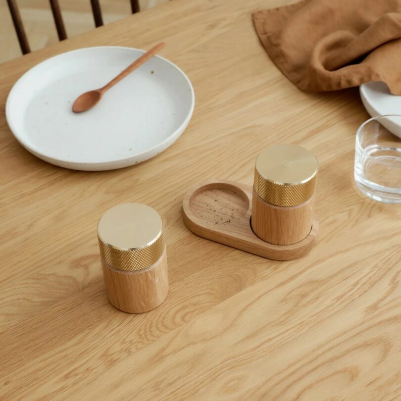 A wooden dining table with a white plate, wooden spoon, brown napkin, glass of water, and a wooden salt and pepper grinder set with gold tops on a small tray