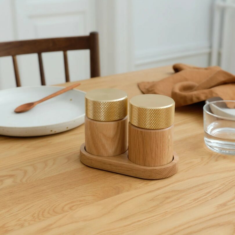 Two wooden containers with gold lids sit on a wooden tray on a dining table, alongside a plate, wooden spoon, glass of water, and brown napkin