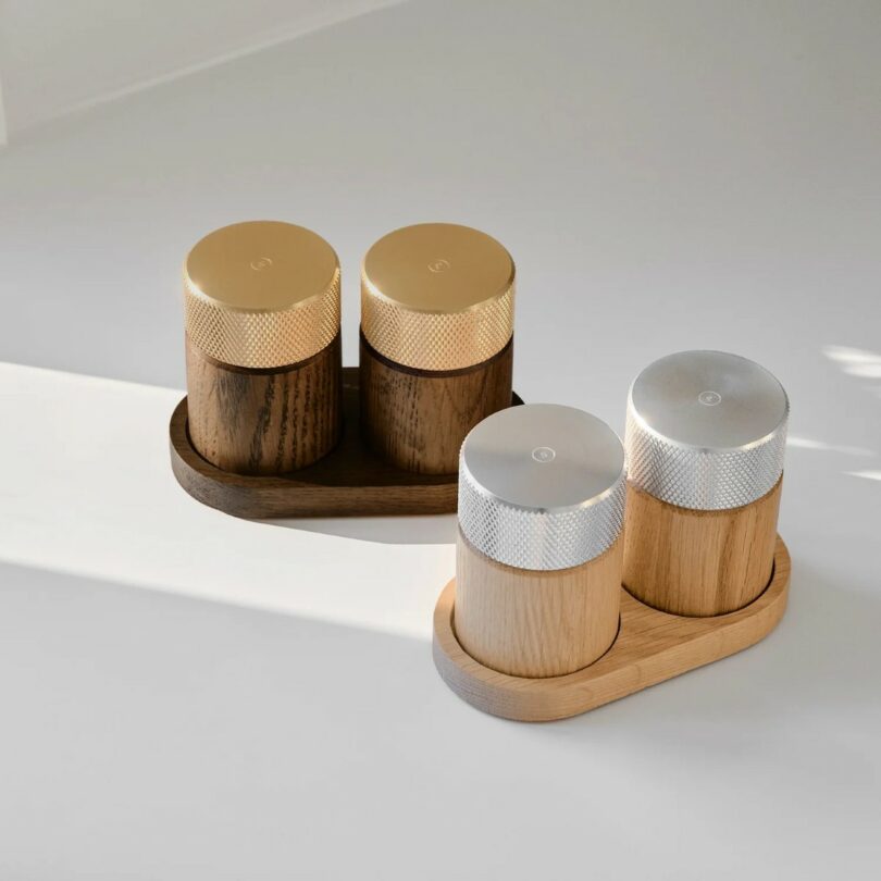 Two sets of wooden salt and pepper shakers with metal tops are displayed on small matching trays, placed on a light surface