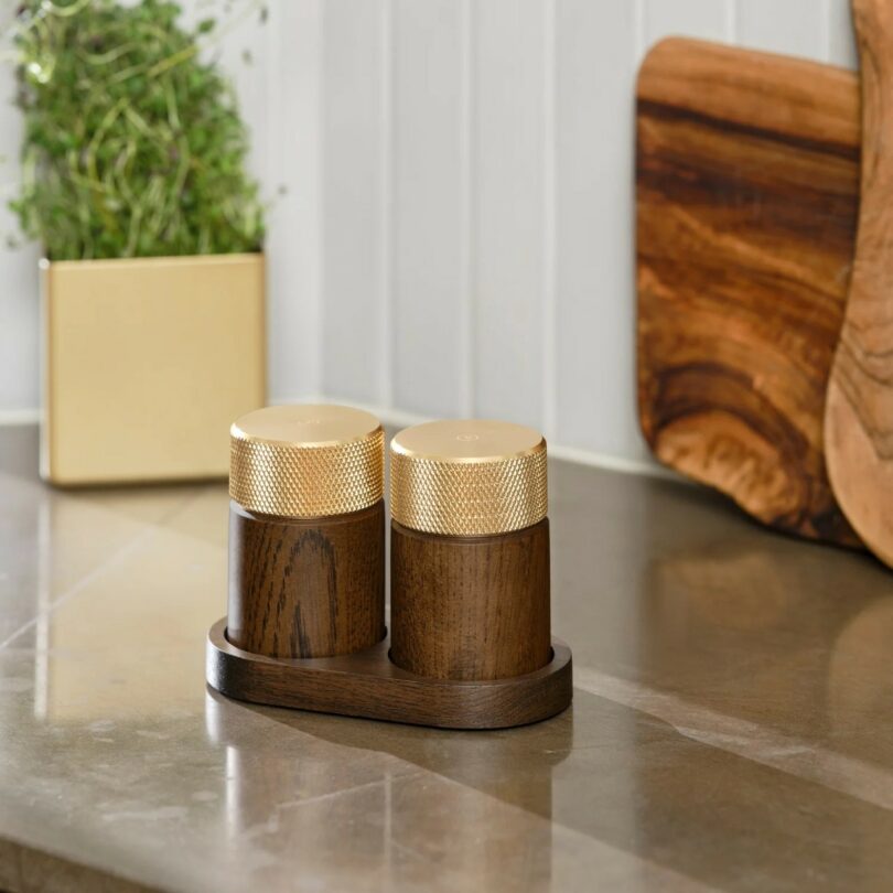 Two wooden spice grinders with gold tops sit on a matching wooden tray on a kitchen counter, with a plant and cutting boards in the background