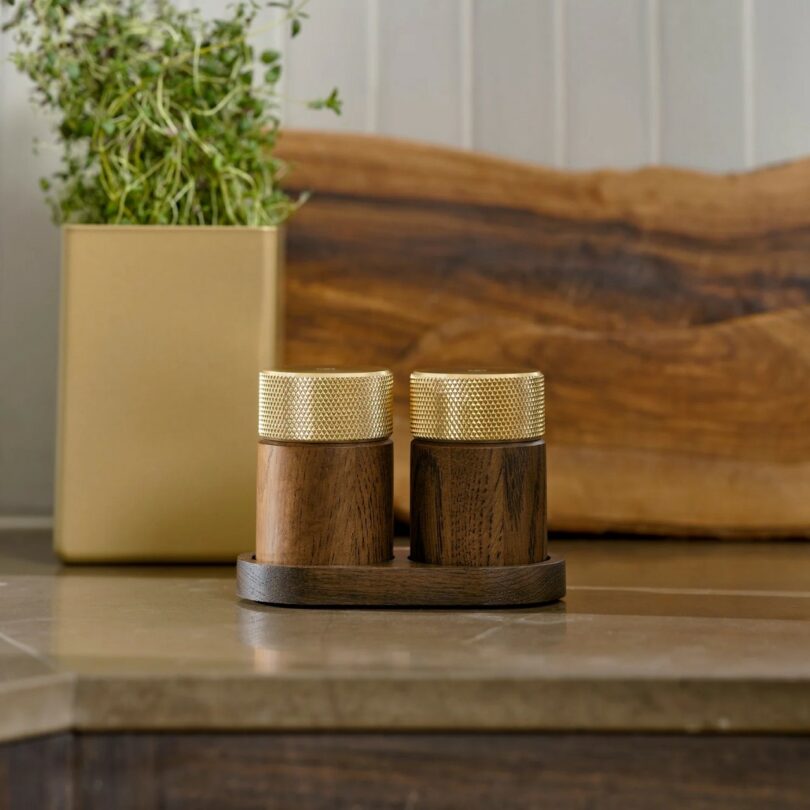 Two wooden containers with textured gold lids sit on a matching wooden tray on a countertop, with a potted plant and wooden cutting board in the background