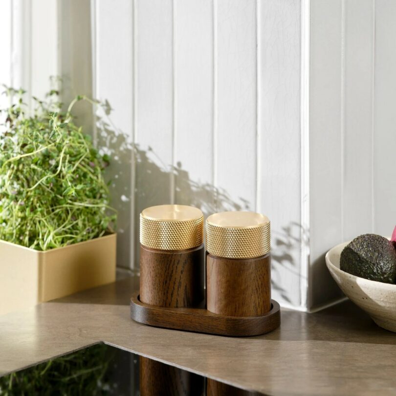 Two wooden salt and pepper grinders with gold tops are placed on a small tray on a kitchen countertop next to a potted herb and a bowl of avocados